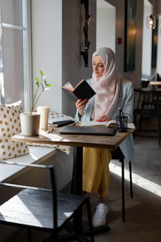 A young woman in a hijab reading a book at a cozy cafe table by the window.
